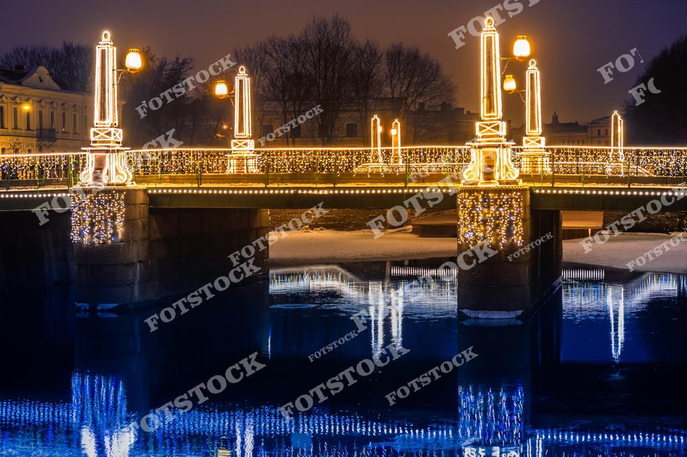 На фотографии изображён мост в Санкт-Петербурге, украшенный к Новому году или Рождеству. Световая иллюминация из гирлянд и фонарей создаёт атмосферу праздника и добавляет сияния тёмной ночи. Отражение огней в воде усиливает эффект, создавая двойную световую композицию. Этот мост, украшенный для зимних праздников, символизирует красоту и уют зимнего Петербурга.